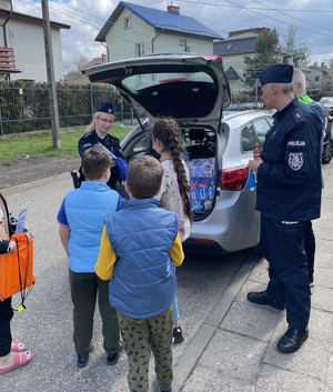 Zdjęcie przedstawia policjanta i policjantkę w granatowych mundurach, którzy stoją przy zaparkowanym radiowozie, którego klapa bagażnika jest otwarta. Obok nich stoją kilkuletni chłopcy.