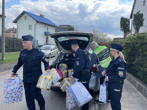 Zdjęcie przedstawia policjantów w mundurach, którzy wyjmują z bagażnika radiowozu kolorowe torby z darami.