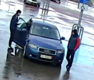 Zdjęcie przedstawia dwóch mężczyzn stojących przy zaparkowanym pojeździe. Jeden z nich przytrzymuje otwarte drzwi od strony pasażera.