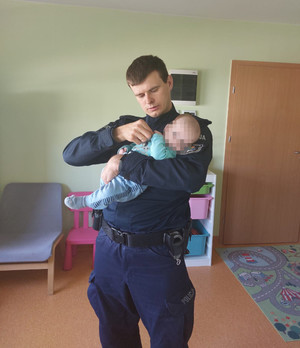 Zdjęcie przedstawia policjanta w granatowym mundurze, który trzyma w ramionach małe dziecko w niebieskich śpioszkach. Twarz dziecko jest zasłonięta komputerowym filtrem.