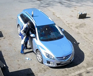 Zdjęcie przedstawia ujęcie z kamery monitoringu pokazujące zaparkowany pojazd m-ki Mazda. Po jego lewej stronie od drzwi pasażera przez otwarte okno do środka sięga mężczyzna. ma on na sobie kolorowy dres.