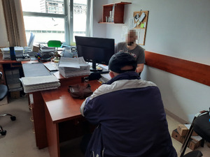 Zdjęcie przedstawia policjanta siedzącego za biurkiem przed ekranem komputera, ma on zasłoniętą twarz filtrem komputerowym. Jest ubrany w szarą koszulkę na krótki rękaw. Przy biurku, tyłem do wejścia i obiektywu aparatu siedzi też zatrzymany mężczyzna. Ma on czarną czapkę na głowie i ubrany jest w kolorowy dres.