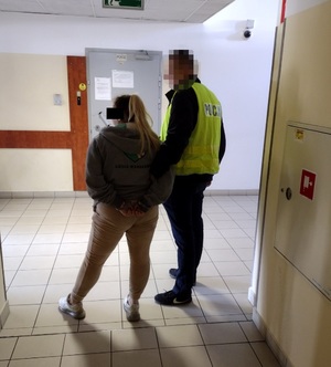 Zdjęcie przedstawia dzielnicowego w odblaskowej kamizelce z napisem POLICJA, który stoi na korytarzu z zatrzymaną kobietą. Ma ona długie włosy związane w kucyk, jest ubrana w brązową bluzę i brązowe spodnie.