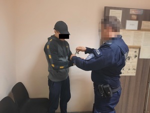 Zdjęcie przedstawia umundurowanego policjanta, który zakłada kajdanki na ręce trzymane z przodu zatrzymanemu mężczyźnie. Policjant jest umundurowany. Jego twarz jest zniekształcona komputerowo. Mężczyzna jest ubrany  w ciemną bluzę z pomarańczowymi wstawkami i ciemne spodnie. Na głowie ma czarną czapeczkę z daszkiem. Część twarzy zasłania mu czarny prostokąt uniemożliwiający jego identyfikację. Znajdują się oni w pomieszczeniu dla osób zatrzymanych.