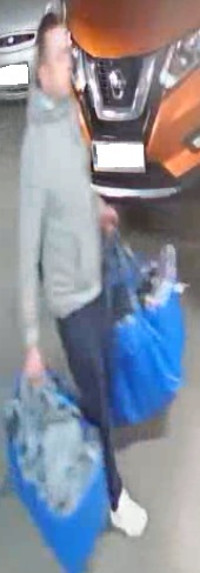 Zdjęcie przedstawia mężczyznę na tle zaparkowanych pojazdów. Rysopis mężczyzny: wiek około 35 lat, około 170 cm wzrostu, krępej budowy ciała, ubrany w szarą bluzę z kapturem, ciemne spodnie, białe buty sportowe. Mężczyzna niesie w obu rękach duże niebieskie torby.