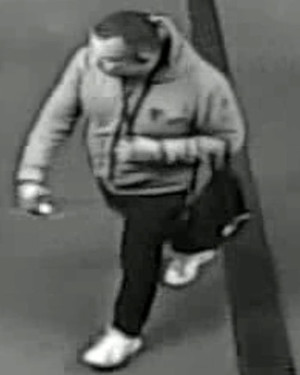 Zdjęcie przedstawia mężczyznę, który ma około 170 cm wzrostu, jest krępej budowy ciała, ubrany w szarą bluzę z kapturem, ciemne spodnie, białe buty sportowe, na lewym przedramieniu niesie czarny plecak.