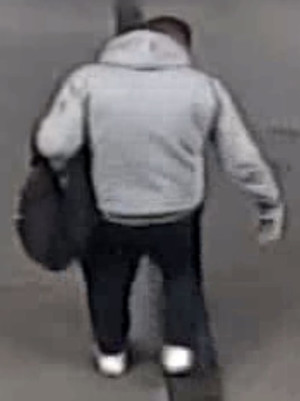 Zdjęcie przedstawia mężczyznę, który ma około 170 cm wzrostu, jest krępej budowy ciała, ubrany w szarą bluzę z kapturem, ciemne spodnie, białe buty sportowe, na lewym przedramieniu niesie czarny plecak. Jest skierowany tyłem do kamery.