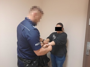 Zdjęcie przedstawia umundurowanego policjanta, który zakłada kajdanki na ręce trzymane z przodu zatrzymanej kobiecie. Policjant jest umundurowany. Jego twarz jest zniekształcona komputerowo. Kobieta ma ciemne włosy związane w kucyk, ubrana jest w ciemną bluzkę i niebieskie spodnie jeansowe. Cześć jej twarzy zasłania czarny prostokąt uniemożliwiający jej identyfikację. Znajdują się oni w pomieszczeniu dla osób zatrzymanych.