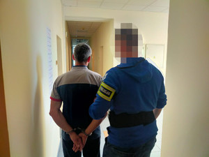 Zdjęcie przedstawia policjanta w ubraniu cywilnym, który prowadzi przez korytarz zatrzymanego mężczyznę. Policjant jest ubrany w niebieską bluzę, na lewym przedramieniu ma odblaskową opaskę z napisem POLICJA. Zatrzymany mężczyzna ma kajdanki na rekach trzymanych z tyłu. Jest ubrany w koszulkę na krótki rękaw. Obaj stoją tyłem do obiektywu aparatu. Twarz i głowa policjanta jest niewyraźna.