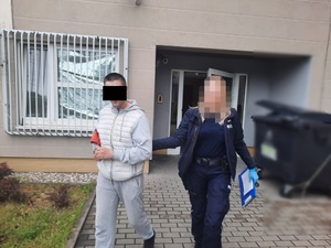 Policjantka w granatowym mundurze prowadzi po dziedzińcu zatrzymanego mężczyznę. Oboje idą na wprost aparatu. Twarz policjantki jest zasłonięta. Większość część twarzy mężczyzny zasłania czarny prostokąt. Jest on ubrany w szary dres. Na rękach trzymanych z przodu ma założone kajdanki. W tle widać budynek komendy i okratowane okno pomieszczenia dla osób zatrzymanych. Policjantka ma na dłoniach niebieskie rękawiczki ochronne, prawą dłonią przytrzymuje prowadzonego mężczyznę za lewe przedramię, w lewej trzyma niebieską teczkę.