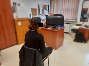 Zdjęcie przedstawia siedzącą w pokoju służbowym na krześle zatrzymaną kobietę. Ma ona ciemne włosy, jest ubrana na czarno. Część jej twarzy zasłania jej czarny prostokąt. Siedzi ona tyłem do obiektywu aparatu. Naprzeciwko niej, za biurkiem za monitorem komputera siedzi policjant. Jego twarz jest zasłonięta komputerowym filtrem.