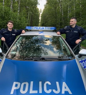 Zdjęcie przedstawia dwóch umundurowanych policjantów, którzy stoją przy oznakowanym radiowozie.