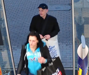 Zdjęcie przedstawia kobietę i mężczyznę wchodzących do sklepu. Kobieta ma ciemne włosy, jest ubrana w rozpiętą ciemną bluzę i niebieską bluzkę. Ma na lewym przedramieniu czarną płócienną torbę z czerwonym napisem. Za nią idzie mężczyzna. Ma on na sobie czarną kurtkę i czarną czapeczkę z daszkiem. W dłoniach trzyma białą reklamówkę, na której widnieje niebieski napis.