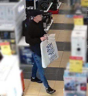 Zdjęcie przedstawia mężczyznę w sklepie. Ma on na sobie czarną kurtkę, niebieskie spodnie jeansowe, czarne buty sportowe i czarną czapeczkę z daszkiem. W dłoniach przed sobą trzyma białą reklamówkę, na której widnieje niebieski napis.
