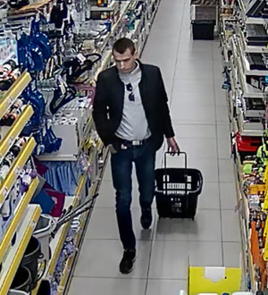 Zdjęcie przedstawia mężczyznę idącego przez sklep. mężczyzna ciągnie za sobą sklepowy wózek. Prawą dłoń trzyma w kieszeni spodni. Jest on ubrany w ciemną kurtkę, jasną bluzkę, ciemne spodnie i ciemne buty, ma ciemne krótkie włosy. Jest frontem do kamery monitoringu.