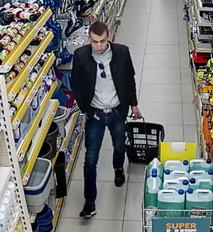 Zdjęcie przedstawia mężczyznę idącego przez sklep. mężczyzna ciągnie za sobą sklepowy wózek. Prawą dłoń trzyma w kieszeni spodni. Jest on ubrany w ciemną kurtkę, jasną bluzkę, ciemne spodnie i ciemne buty, ma ciemne krótkie włosy. Jest frontem do kamery monitoringu.