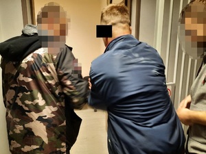 Zdjęcie przedstawia dwóch policjantów w ubraniach cywilnych - jeden ma na sobie kurtkę we wzory, drugi szarą bluzę. Pomiędzy nimi znajduje się zatrzymany mężczyzna. Ma on na sobie niebieską kurtkę. Wszyscy wchodzą do pomieszczenia dla osób zatrzymanych. Stoją tyłem do obiektywu aparatu. Zatrzymany mężczyzna ma część twarzy zasłoniętą czarnym prostokątem. Twarze policjantów są zniekształcone komputerowym filtrem.