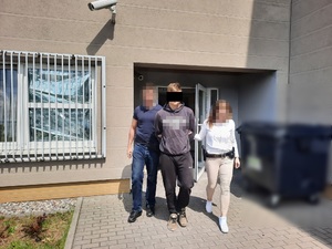 Zdjęcie przedstawia policjanta i policjantkę w ubraniach cywilnych, którzy wyprowadzają z budynku zatrzymanego mężczyznę. Mężczyzna ten znajduje się pomiędzy policjantami. Trzyma ręce z tyłu. Część jego twarzy zasłania czarny prostokąt. Jest ubrany w szarą bluzę i szare spodnie. Twarze policjantów są zamazane.