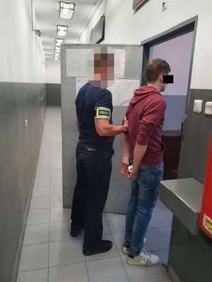 Zdjęcie przedstawia policjanta, który wprowadza do celi zatrzymanego mężczyznę. Policjant przytrzymuje za lewe przedramię stojącego po jego prawej stronie młodego mężczyznę. Na prawym przedramieniu policjant ma odblaskową opaskę z napisem POLICJA. Zatrzymany mężczyzna ma na sobie czerwoną bluzę i niebieskie spodnie jeansowe. Drzwi do celi są otwarte.