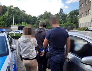 Zdjęcie przedstawia policjantów w ubraniach cywilnych, którzy prowadzą zatrzymanego mężczyznę. Ma on kajdanki założone na rękach trzymanych z tyłu. Wszyscy stoją pomiędzy radiowozem i innym cywilnym samochodem. Stoją tyłem do obiektywu aparatu. W tle widać inne zaparkowane pojazdy.