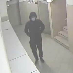 Zdjęcie przedstawia mężczyznę idącego korytarzem bloku. Ma on około 180 cm wzrostu, ubrany jest w ciemną kurtkę, ciemne spodnie, czapkę z daszkiem, twarz zasłania mu maseczka ochronna.