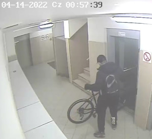 Zdjęcie przedstawia mężczyznę, który stoi z rowerem przy windzie. Ma on około 180 cm wzrostu, ubrany jest w ciemną kurtkę, ciemne spodnie (z tyłu na prawej nogawce widoczny biały napis „P56”), czapkę z daszkiem, na plecach ma plecak z symbolem NIKE. Stoi tyłem do kamery monitoringu.