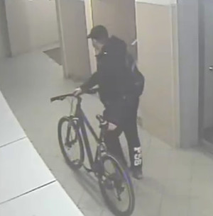 Zdjęcie przedstawia mężczyznę, który stoi z rowerem na korytarzu. Ma on około 180 cm wzrostu, ubrany jest w ciemną kurtkę, ciemne spodnie (z tyłu na prawej nogawce widoczny biały napis „P56”), czapkę z daszkiem, na plecach ma plecak z symbolem NIKE. Stoi bokiem do kamery monitoringu. W tle widać drzwi do windy.