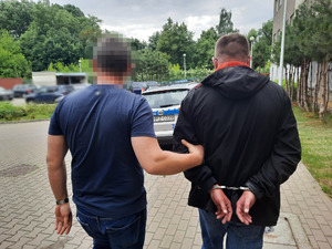 Zdjęcie przedstawia policjanta ubranego w granatową koszulkę na krótki rękaw i granatowe spodnie jeansowe, który prowadzi zatrzymanego mężczyznę, przytrzymując go za lewe przedramię. Mężczyzna ten ma na sobie ciemną kurtkę i granatowe spodnie jeansowe. Na rękach trzymanych z tyłu ma założone kajdanki. Obaj stoją tyłem do obiektywu aparatu. W tle widać zaparkowane pojazdy.