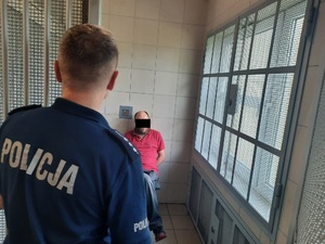 Zdjęcie przedstawia siedzącego w celi zatrzymanego mężczyznę. Jest on ubrany w czerwoną koszulkę na krótki rękaw. Część twarzy zasłania mu czarny prostokąt. Siedzi podparty o ścianę, przodem do wejścia i do obiektywu aparatu. W otwartych drzwiach stoi policjant w granatowym mundurze. Stoi on tyłem do obiektywu aparatu i przodem do mężczyzny.