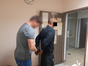 Zdjęcie przedstawia policjanta ubranego w szarą koszulkę na krótki rękaw i niebieskie spodnie jeansowe, który zakłada kajdanki zatrzymanemu mężczyźnie na ręce trzymane z tyłu. Policjant ma na dłoniach ciemne rękawiczki. Zatrzymany mężczyzna jest ubrany w ciemną bluzę na długi rękaw i ciemne spodnie. Obaj znajdują się w pomieszczeniu dla osób zatrzymanych. Stoją prawym profilem do obiektywu aparatu. W tle widać informacje umieszczony na drzwiach pomieszczenia i okratowane drzwi prowadzące do innej części pomieszczenia.