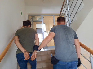 Zdjęcie przedstawia policjanta ubranego w szarą koszulkę na krótki rękaw i niebieskie spodnie jeansowe, który prowadzi zatrzymanego mężczyznę. Policjant przytrzymuje idącego po jego lewej stronie mężczyznę za spojenie kajdanek, które ten ma założone na rękach trzymanych z tyłu. Policjant ma na dłoniach ciemne rękawiczki. Zatrzymany mężczyzna jest ubrany w ciemną bluzę na długi rękaw i ciemne spodnie. Obaj znajdują się tyłem do obiektywu aparatu. W tle widać oszklone drzwi prowadzące na korytarz.