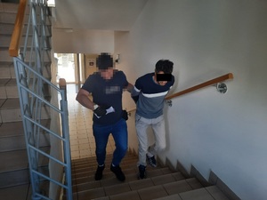 Zdjęcie przedstawia policjanta ubranego w granatową koszulkę na krótki rękaw i niebieskie spodnie jeansowe, który wprowadza po schodach zatrzymanego mężczyznę. Policjant ma zasłoniętą twarz komputerowym filtrem, na dłoniach ma czarne rękawiczki, lewą ręką przytrzymuje idącego po jego lewej stronie mężczyznę. Mężczyzna ten ma sweter w kolorze granatowo-szare i białe pasy oraz jasnoniebieskie spodnie jeansowe. Na rękach trzymanych z tyłu ma założone kajdanki. Część twarzy zasłania mu czarny prostokąt. Obaj znajdują się przodem do obiektywu aparatu.
