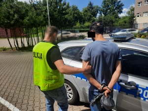 Zdjęcie przedstawia policjanta ubranego w koszulkę na krótki rękaw w kolorze ciemnym i seledynowy bezrękawnik z napisem POLICJA, który prowadzi zatrzymanego mężczyznę do zaparkowanego oznakowanego radiowozu. Twarz policjanta jest ukryta pod komputerowym filtrem. Zatrzymany mężczyzna ten ma ciemne włosy i ciemną karnację. Jest ubrany w grafitową koszulkę na krótki rękaw i niebieskie spodnie jeansowe. Część twarzy zasłania mu czarny prostokąt. Na rękach trzymanych z tyłu ma założone kajdanki. Obaj znajdują się tyłem do obiektywu aparatu. W tle widać zaparkowane pojazdy.