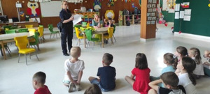 Zdjęcie przedstawia umundurowanego policjant, który stoi w sali przed siedzącymi na dywanie dziećmi. W tle, na ścianach widać kolorowe prace i rysunki.