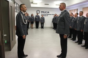 Zdjęcie przedstawia dwóch funkcjonariuszy stojących naprzeciwko siebie w sali w uroczystych mundurach. W tle widać innych zebranych w sali policjantów ubranych w galowe mundury.