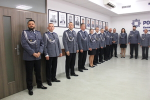 Zdjęcie przedstawia funkcjonariuszy w uroczystych mundurach stojących w sali w jednym szeregu. Nad nimi widać powieszone na ścianie portrety osób umieszczone w ramach.