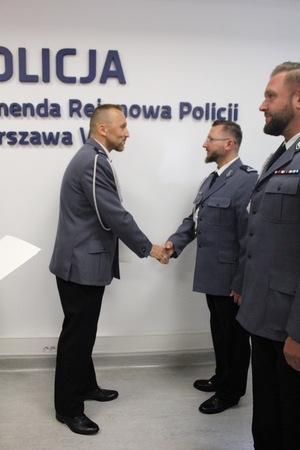 Zdjęcie przedstawia funkcjonariuszy stojących naprzeciwko siebie i podających sobie ręce, są oni ubrani w uroczyste mundury.