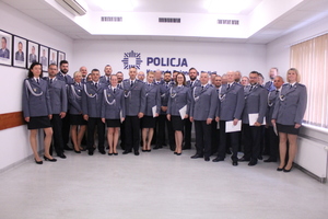 Zdjęcie przedstawia funkcjonariuszy stojących pod ścianą, na której widnieje napis KOMENDA REJONOWA POLICJI WARSZAWA VI, są oni ubrani w uroczyste mundury. Wszyscy uśmiechają się do obiektywu aparatu.