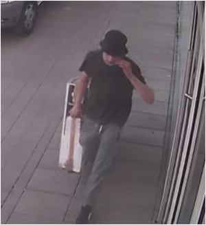 Zdjęcie przedstawia postać młodego mężczyzny w wieku około 20-30, wzrostu około 185 cm lat idącego chodnikiem. Ma on na sobie czarną koszulkę na krótki rękaw, szare spodnie i czarne bytu z białymi elementami. Na głowie ma ciemny kapelusz. W prawej ręce trzyma prostokątny karton. Lewą naciąga kapelusz na oczy. W tle widać fragment samochodowego koła.