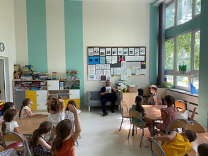 Zdjęcie przedstawia salę szkolną, a w niej przy okrągłych stolikach siedzą mali uczniowie. Przodem do nich, na kanapie pod ścianą siedzi umundurowany policjant. Trzyma on kolorową książeczkę w ręce.