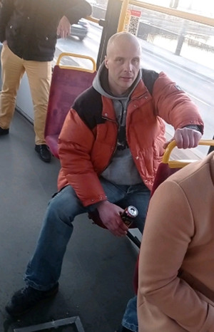 Zdjęcie przedstawia mężczyznę siedzącego na krześle w tramwaju. W prawej dłoni trzyma jakaś puszkę z napojem. Patrzy na wprost kamery. Za nim widać stojącego innego pasażera. Mężczyzna ma na sobie czerwoną kurtkę puchową, pod nią szarą bluzę, niebieskie spodnie jeansowe i czarne buty.