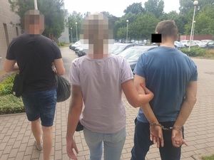 Zdjęcie przedstawia policjantkę ubraną w jasną koszulkę na krótki rękaw i jasnoniebieskie spodnie jeansowe, która prowadzi po swojej prawej stronie zatrzymanego mężczyznę. Policjantka przytrzymuje mężczyznę za lewe przedramię. Na rękach trzymanych z tyłu mężczyzna ma założone kajdanki. Mężczyzna jest ubrany w granatową koszulkę na krótki rękaw i ciemne długie spodnie. Część lewego profilu zasłania mu czarny prostokąt. Przed idącą policjantką, idzie inny policjant ubrany po cywilnemu w ciemną koszulkę na krótki rękaw i ciemne spodenki o długości do kolan. niesie on w rekach czarny worek z zawartością. Wszyscy są tyłem do obiektywu aparatu. Po ich lewej stronie widać ścianę budynku komendy, a przed nimi w tle są zaparkowane pojazdy.