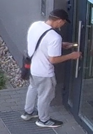 Zdjęcie przedstawia mężczyznę, który próbuje otworzyć drzwi budynku. Ma około 175 cm wzrostu. Ubrany jest w białą koszulkę na krótki rękaw, szare spodnie, czarne buty sportowe z białym paskiem przy podeszwie, czarną czapkę z daszkiem; przez ramię ma przewieszoną torebkę koloru czarno-czerwonego.