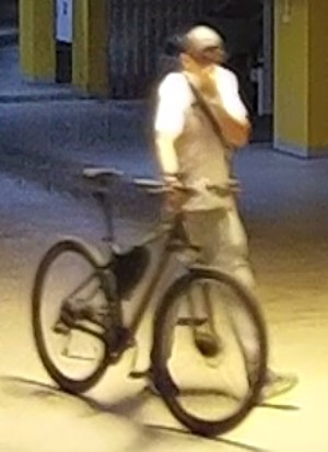 Zdjęcie przedstawia sylwetkę mężczyzny prowadzącego rower. Ubrany jest w jasną koszulkę i spodnie, ciemne buty, czapkę z daszkiem.