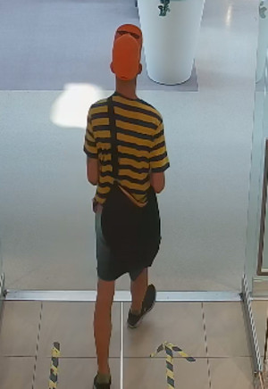 Zdjęcie przedstawia szczupłą sylwetkę mężczyzny, ubrany on jest w jasnopomarańczową koszulkę z krótkim rękawem w czarne poziome paski, krótkie spodenki typu jeans, pomarańczową czapkę z daszkiem na której ma założone okulary przeciwsłoneczne; przez lewe ramię ma przewieszoną czarną torbę. Znajduje się tyłem do kamery monitoringu.