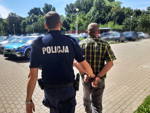 Zdjęcie przedstawia umundurowanego policjanta, który prowadzi przez dziedziniec, po swojej prawej stronie zatrzymanego mężczyznę przytrzymując go za lewe przedramię. Obaj znajdują się tyłem do obiektywu aparatu. Zatrzymany mężczyzna jest ubrany w koszulkę na krótki rękaw w kolorze czarno-żółtym i szare długie spodnie. Na rękach trzymanych z tyłu ma założone kajdanki. W tle widać zaparkowane na dziedzińcu pojazdy oznakowane i nieoznakowane.