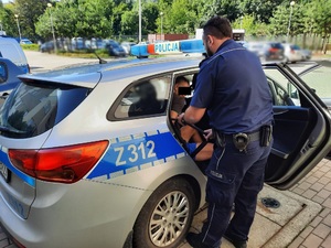 Zdjęcie przedstawia umundurowanego policjanta, który nachyla się nad mężczyzną siedzącym w radiowozie. Mężczyzna ma na sobie niebieskie krótkie spodenki i czarną koszulkę polo z krótkim rękawem, z zieloną obwódką wokół rękawa. Część jego twarzy, w okolicy oczu jest zasłonięta czarnym prostokątem.