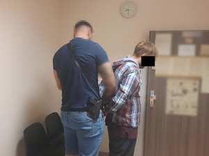 Zdjęcie przedstawia policjanta ubranego w ciemnogranatową koszulkę na krótki rękaw i niebieskie spodenki jeansowe o długości do kolan, który zakłada kajdanki na ręce trzymane z tyłu zatrzymanemu mężczyźnie. Policjant na prawym boku ma czarną kaburę z bronią służbową. Obaj stoją prawym profilem do obiektywu aparatu. Twarz policjanta jest rozmazana. Mężczyzna jest ubrany w koszulę na długi rękaw w kratę czerwono-niebieską, pod nią ma bluzę bordową i ciemne spodnie. Jego twarz jest zasłonięta czarnym prostokątem. W tle widać otwarte drzwi pomieszczenia, na którym są wywieszone kartki z różnymi informacjami.