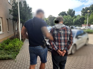 Zdjęcie przedstawia policjanta ubranego w ciemnogranatową koszulkę na krótki rękaw i niebieskie spodenki jeansowe o długości do kolan, który prowadzi po dziedzińcu zatrzymanego mężczyznę. Policjant na prawym boku ma czarną kaburę z bronią służbową. Obaj stoją tyłem do obiektywu aparatu. Twarz policjanta jest zamazana. Mężczyzna jest ubrany w koszulę na długi rękaw w kratę czerwono-niebieską, pod nią ma bluzę bordową i ciemne spodnie. Fragment jego twarzy jest zasłonięta czarnym prostokątem. W tle widać zaparkowane pojazdy.