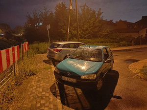 Zdjęcie przedstawia zaparkowany częściowo na ulicy i na chodniku niebieski pojazd m-ki Peugeot, za nim widać srebrny nieoznakowany pojazd policjantów. Zdjęcie jest robione wieczorem. W tle widać ogrodzenia posesji.